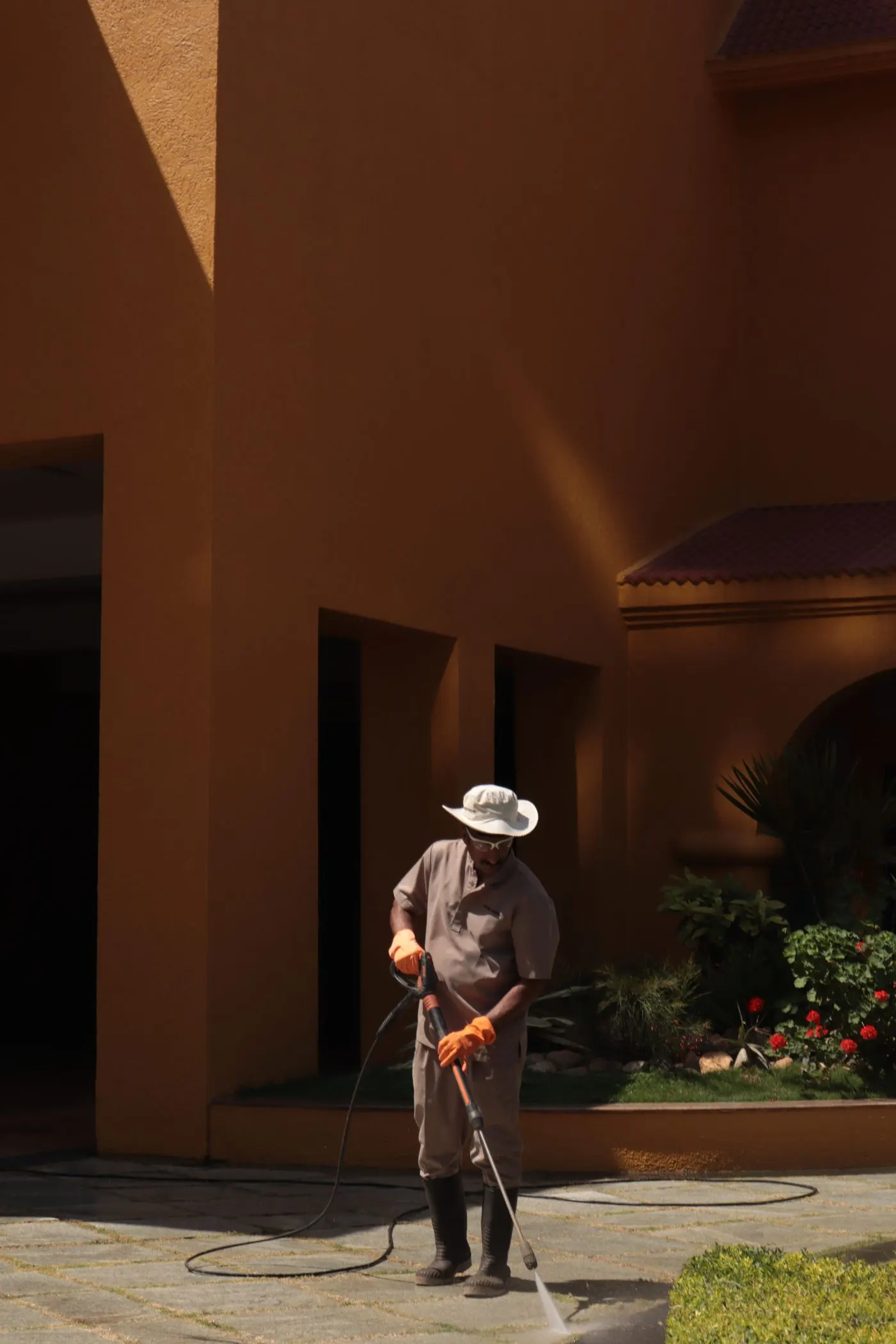 Worker in a sun hat pressure washing stone paving beside a warm orange stucco building