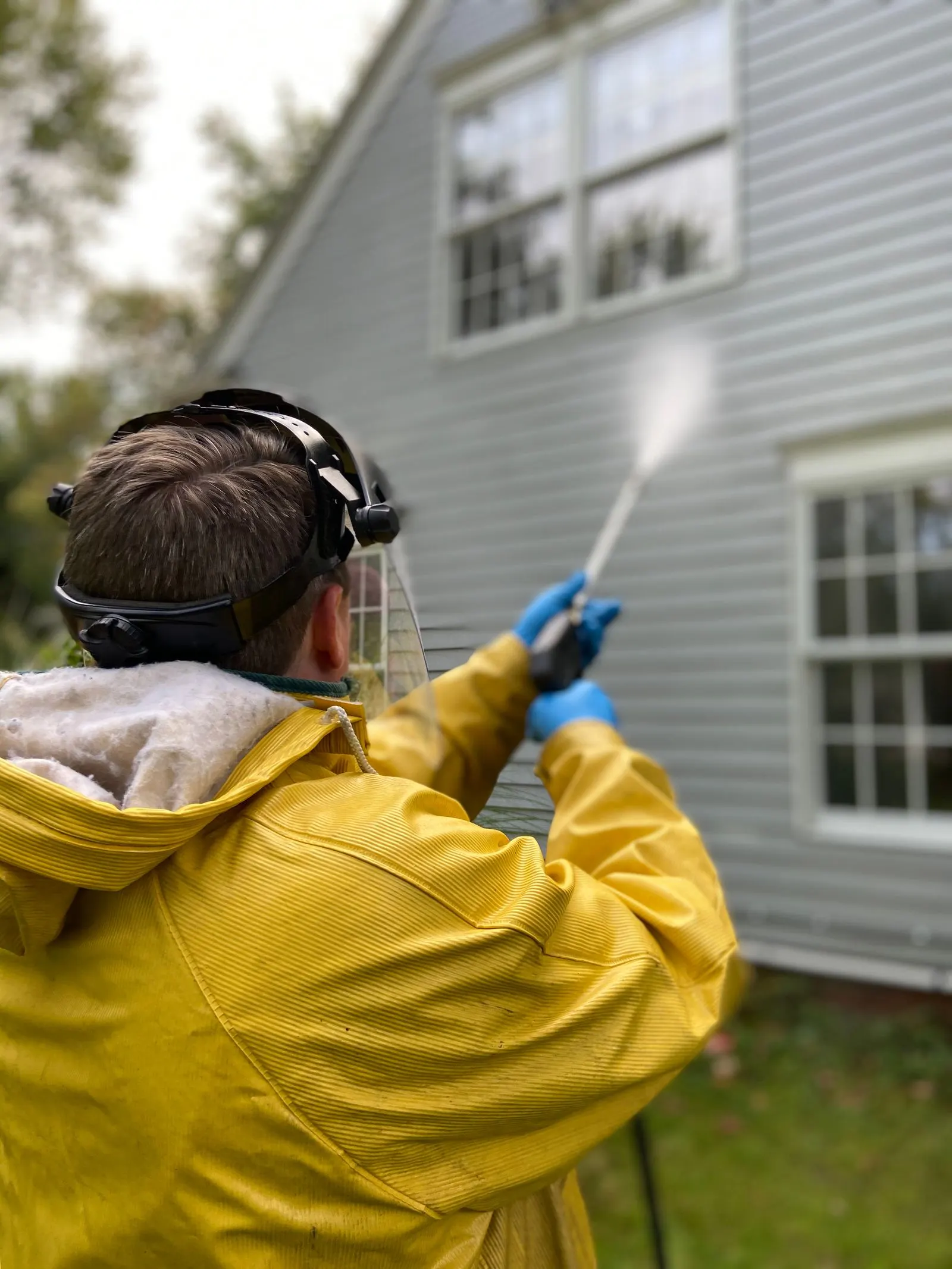 Person in a yellow rain jacket and face shield spraying the siding of a gray house with a wand